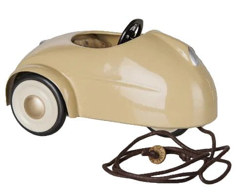 A vintage-style beige pedal toy car with black wheels, a black steering wheel, and a brown coiled cord with a round switch, shown on a gray checkered background.