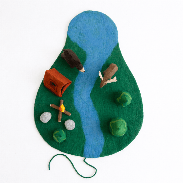 A felt play mat shaped like a landscape with a blue river winding through green hills, decorated with small toy animals, a red-orange tent, a wooden cross with a yellow flame, and green and gray stones, photographed on a white background.