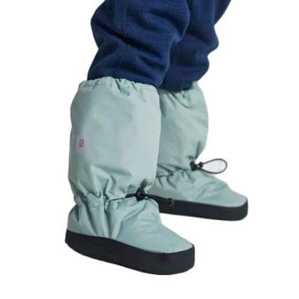 A child wearing dusty mint green snow boots with black soles and black drawstrings, paired with navy fleece pants, shown from the side against a light gray studio background.