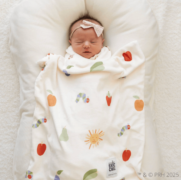 A sleeping newborn baby swaddled in a cream-colored blanket with colorful fruit and caterpillar prints, lying on a white U-shaped bassinet. The blanket features illustrations of apples, pears, a sun, and caterpillars.