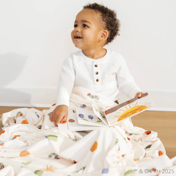 Child sitting on a bed with a colorful book, surrounded by a white blanket with fruit patterns.