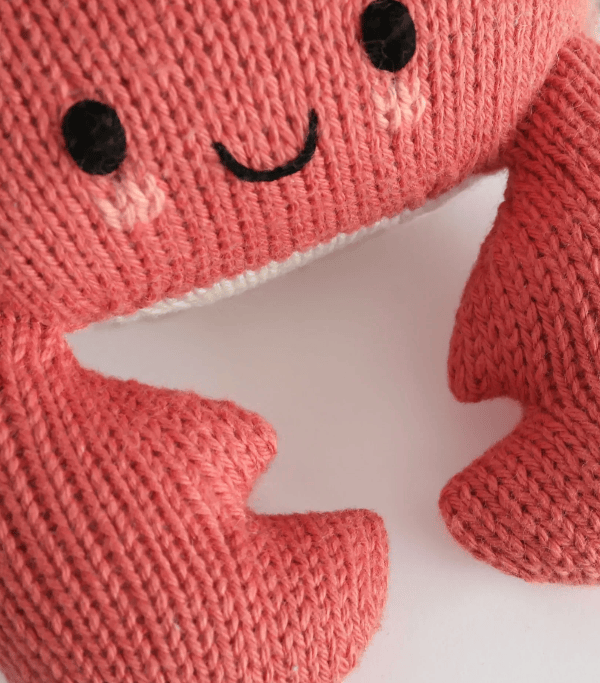 Close-up of a knitted plush crab toy in a coral pink color, featuring embroidered black eyes and a smile on a white background.