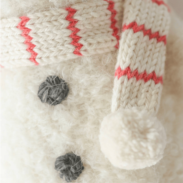 Close-up of a soft knit snowman accessory, likely a hat or scarf, in off-white with red zigzag stripes and a fluffy pom-pom, shown against a white fuzzy background with dark buttons.