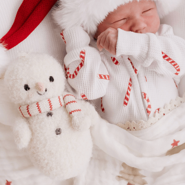 A sleeping newborn baby wearing a white knit holiday romper with red candy cane patterns, a white faux-fur hat, and a red Santa hat, lying next to a plush white snowman toy on a soft white blanket.