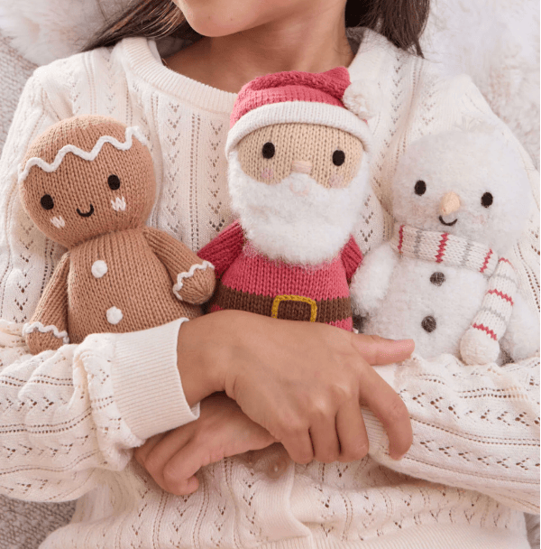 A person wearing a cream cable-knit sweater holds three handmade knitted holiday toys: a gingerbread man, a Santa Claus figure, and a white snowman with a striped scarf.