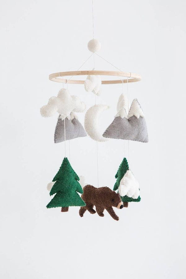 A felt baby mobile hanging from a light wood ring, featuring white clouds, gray mountains, a white crescent moon, green pine trees, and a brown bear on a white background.