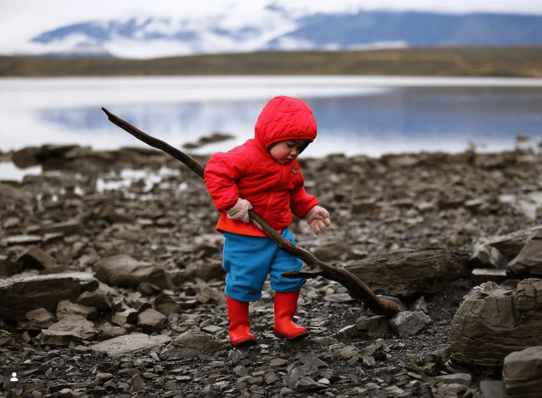 A toddler wearing a bright red hooded jacket, blue pants, and red rain boots stands on a rocky lakeshore holding a long dark branch, with water and mountains in the background.