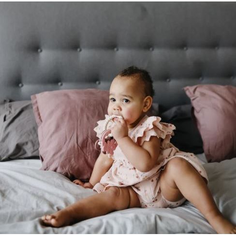 Baby in a pink dress sitting on a bed with pink pillows and a gray headboard.